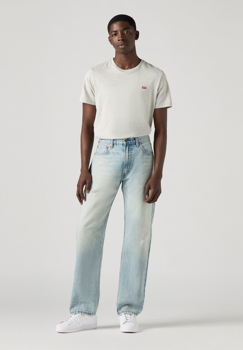 T-shirt texturé gris clair avec un petit logo rouge, associé à un jean déchiré bleu clair taille haute et des baskets blanches.