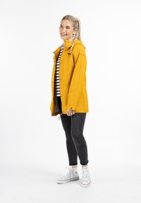 Giacca impermeabile gialla con fodera bianca, cappuccio regolabile e due tasche frontali. Abbinata a una maglietta a righe, jeans neri e sneakers bianche.