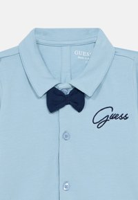 Ανοιχτό μπλε πόλο πουκάμισο με μπλε-σκούρο παπιγιόν, κεντημένο λογότυπο "Guess" και κλείσιμο με τρία κουμπιά. Υλικό από απαλό βαμβάκι.