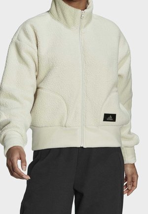 Osoba nosząca białą, puszystą kurtkę zapinaną na zamek z wysokim kołnierzem oraz czarne spodnie, z naszywką z logo Adidas na przedniej części kurtki.
