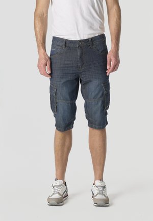 Hombre vestido con pantalones cortos de mezclilla azul marino hasta la rodilla con múltiples bolsillos, zapatillas blancas y camiseta blanca, de pie sobre un fondo liso.