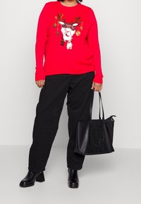 Persona che indossa un maglione rosso con una renna di paillettes e pantaloni neri, tiene in mano una borsa a tracolla in pelle nera e indossa stivali neri con tacco.