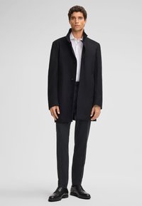 Cappotto in misto lana nero con colletto alto, chiusura con bottoni e due tasche frontali, abbinato a pantaloni scuri e scarpe nere.
