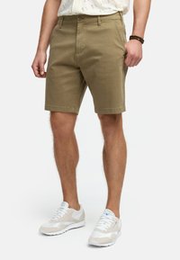 Khaki-shortsit, jotka on valmistettu puuvillasta, klassisella istuvuudella, kahdella etutaskulla ja nappisuljennalla. Yhdistetty vaaleanvärisiin urheilujalkineisiin.