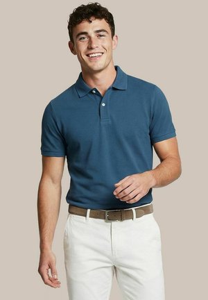 Jonge man die lacht, draagt een blauw poloshirt met korte mouwen, witte broek en een bruine riem, staand tegen een effen beige achtergrond.