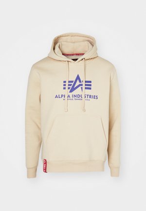 Bež hoodie s velikim ljubičastim logotipom i dizajnerskim detaljima "Alpha Industries". Uključuje prednju džep i crvenu oznaku na rubu.