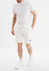 Witte katoenen shorts met een drawstring taille, zijzakken en een blauw "Creeks" logo op het linkerpoot, gedragen met een plain wit T-shirt.