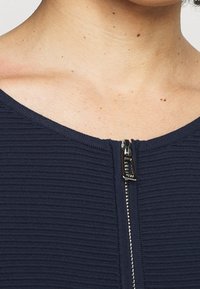 Granatowy top z prążkowanej tkaniny z centralnym zamkiem błyskawicznym, metalowymi elementami i szerokim dekoltem. Tekstura jest gładka z delikatnym rozciągnięciem.