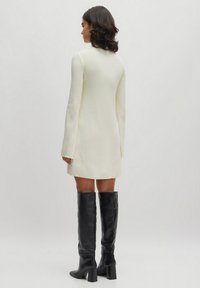 Weißes Strickkleid mit langen Ärmeln und schwingendem Saum, kombiniert mit hohen schwarzen Lederstiefeln mit einem quadratischen Absatz. Minimalistisches Design.