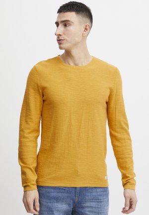 Długoobranżowa koszulka crew neck w kolorze musztardowym. Wykonana z miękkiego, strukturalnego materiału o luźnym kroju. Posiada małą metkę z logo przy dolnym brzeg.