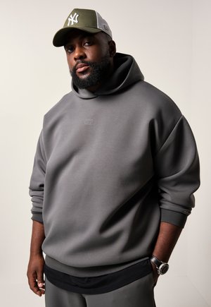 Grauer Kapuzenpullover mit Rippbündchen, versehen mit einem kleinen Logo auf der Brust. Eine grüne Mütze mit einem weißen Logo rundet das Outfit ab.