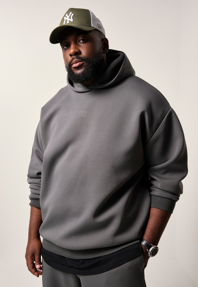 Grijze sweatshirt met een capuchon en ribgebreide manchetten, met een klein logo op de borst. Een groene pet met een wit logo maakt de look compleet.