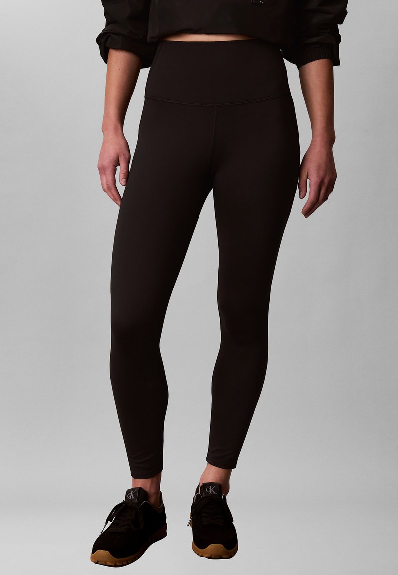Femme portant des leggings noirs taille haute et des baskets noires avec des semelles beige, debout devant un fond gris uni.