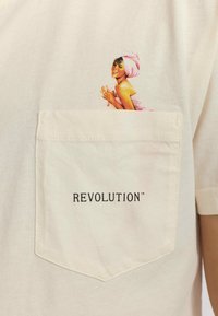 Camiseta color crema con un bolsillo frontal que lleva la palabra "REVOLUCIÓN™" en negro. El bolsillo muestra un gráfico colorido de una mujer.