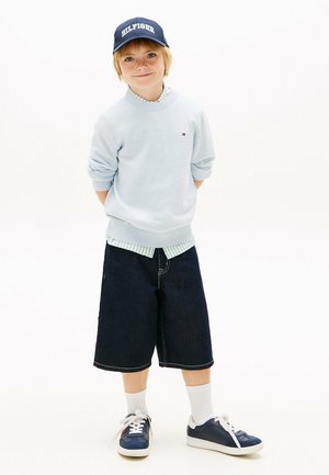 Jonge jongen met een licht truitje, donkere denim shorts, een marineblauwe pet en sneakers, staand met handen achter de rug tegen een witte achtergrond.