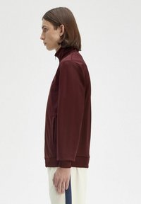 Giacca a zip color bordeaux con colletto alto e tasche laterali. Tessuto liscio, design aderente e polsini a coste. Modello mostrato di lato.