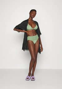 Kimono de renda preta sobre um conjunto de soutiens e cuecas de cintura alta com acabamento em renda verde, combinado com sandálias deslizantes lavanda.