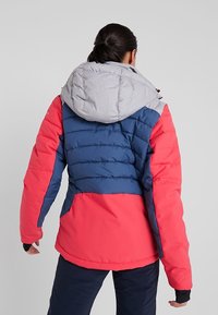 Gepolsterte Jacke mit grauer Kapuze, blauem Mittelteil und roten Ärmeln. Strukturierter Stoff mit horizontalen Steppnähten zur Isolierung.