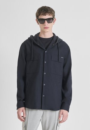 Marineblauw button-up jas met een capuchon, twee borstzakken en een gestructureerde afwerking. G gedragen over een zwart shirt, gecombineerd met grijze broek en zonnebril.