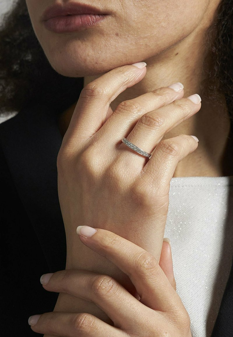 Silberner Ring mit strukturiertem Oberflächen und schmalem Band, getragen an einer Hand mit manikürten Nägeln, vor einem dunklen Hintergrund.