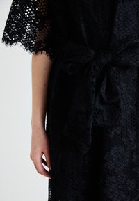 Vêtement en dentelle noire avec des motifs découpés complexes, manches courtes et une ceinture nouée à la taille. Texture douce.