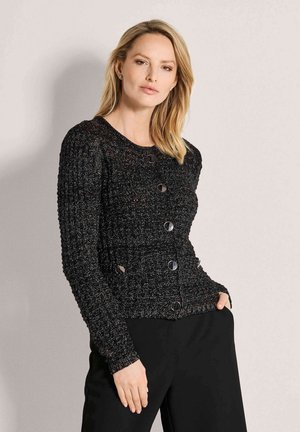 Blonde Frau, die einen schwarzen, strukturierten Knopf-Pullover und schwarze Hosen trägt, steht mit einer Hand in der Tasche vor hellem Hintergrund.