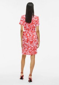 Abito floreale rosso con maniche corte, vita cintrata, orlo all'altezza del ginocchio e tessuto morbido. Indossato con tacchi alti rossi con cinturini.