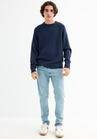 Suéter de punto de color navy con dobladillos y puños acanalados, combinado con jeans azul claro y zapatillas blancas con detalles en marrón. Conjunto casual.