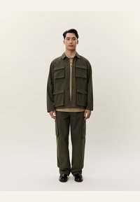 Giovane uomo in piedi con giacca e pantaloni cargo verde oliva, camicia beige e scarpe nere contro uno sfondo chiaro e uniforme.