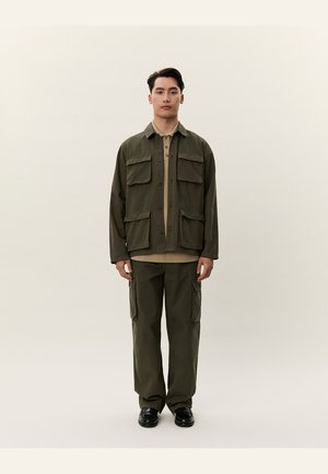 Giovane uomo in piedi con giacca e pantaloni cargo verde oliva, camicia beige e scarpe nere contro uno sfondo chiaro e uniforme.