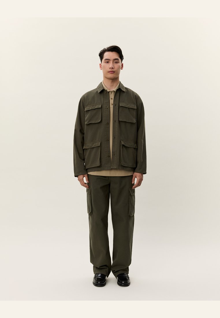 Giovane uomo in piedi con giacca e pantaloni cargo verde oliva, camicia beige e scarpe nere contro uno sfondo chiaro e uniforme.