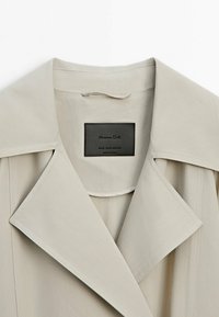 Jasno beżowy trencz z szerokim kołnierzem, wykonany z teksturowanej tkaniny. Posiada czarną metkę z napisem "Massimo Dutti" oraz instrukcje pielęgnacji.