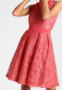 En korallfärgad klänning i spetsdesign med en åtsittande överdel och utsvängd kjol, som har ett solidt midjeband och intrikata blommönster genom hela klänningen.