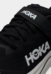 Κοντινό πλάνο μαύρου παπουτσιού τρεξίματος HOKA με μαύρα κορδόνια, ιμάντα με βέλκρο και λευκό λογότυπο στο πλάι και στον ιμάντα, με λευκή σόλα που έχει μαύρη ρίγα.