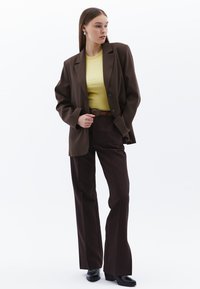 Brun kavaj över en gul topp, matchad med mörkbruna byxor. Inkluderar ett brunt bälte och svarta klackskor. Släta texturer, skräddarsydd passform.