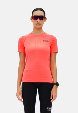 Frau trägt korallenrotes, kurzärmeliges Sportshirt, schwarze Kompressionsleggings, Sportsonnenbrille und eine schwarze Armbanduhr, steht vor weißem Hintergrund.