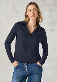 Maglia a maniche lunghe blu navy con colletto e mezza pattina; superficie liscia abbinata a jeans blu casual con dettagli a bottoni.