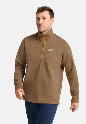 Uomo che indossa un pile marrone con mezza cerniera e collo alto e pantaloni blu scuro, cammina e guarda leggermente di lato.