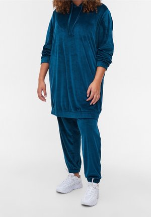 Personne portant un sweat à capuche ample en velours bleu et un pantalon de jogging assorti, associés à des baskets blanches de sport sur un fond uni.