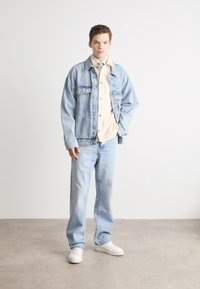 Lichtblauwe spijkerjack over een beige overhemd met knopen, gecombineerd met lichtblauwe spijkerbroek en witte sneakers. Glad textuur, relaxte pasvorm.