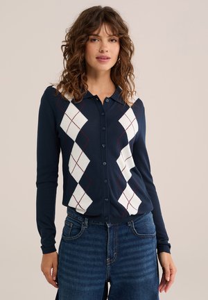 Frau trägt einen marineblauen, geknöpften Cardigan mit weißem und rotem Rautenmuster, kombiniert mit hoch taillierten blauen Jeans, stehend vor beigem Hintergrund.