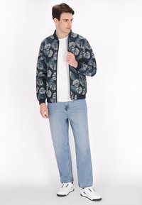 Blomstret bomberjakke i marineblå med hvide bladdekor, ribbede manchetter og kant. Båret over en hvid t-shirt, parret med lyseblå jeans og hvide sneakers.