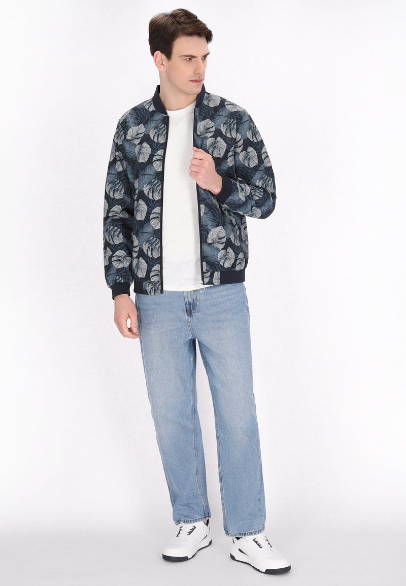 Blomstret bomberjakke i marineblå med hvide bladdekor, ribbede manchetter og kant. Båret over en hvid t-shirt, parret med lyseblå jeans og hvide sneakers.