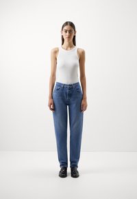 Hvit ermløs tanktopp kombinert med blå straight-leg jeans, med frontlommer og knapplukking. Svarte sko fullfører antrekket.