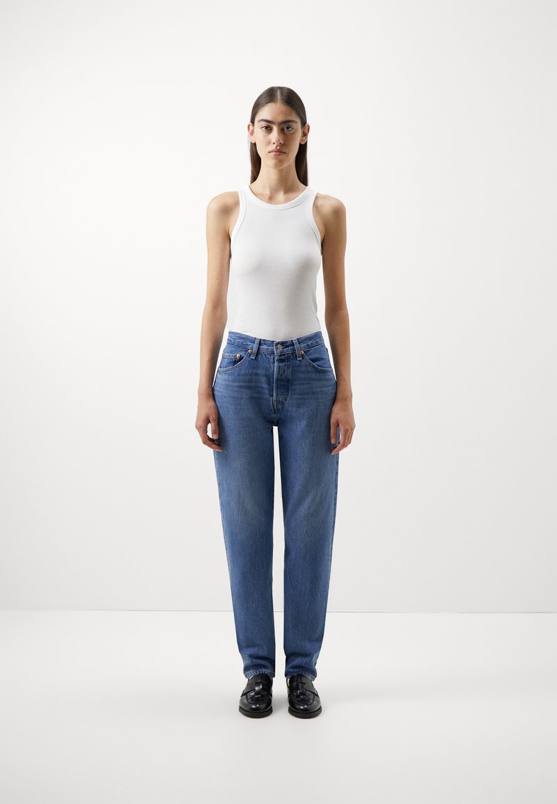 Hvit ermløs tanktopp kombinert med blå straight-leg jeans, med frontlommer og knapplukking. Svarte sko fullfører antrekket.
