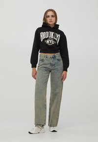 Felpa nera corta con stampa "BROOKLYN NYC", jeans a vita alta blu chiaro e sneakers bianche. Tessuto morbido, vestibilità casual.