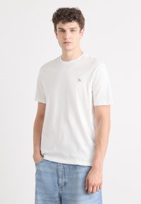 Camiseta de algodón blanca con cuello redondo y mangas cortas. Presenta un pequeño parche de logo rojo en el lado izquierdo del pecho. Combinada con jeans de color azul claro.