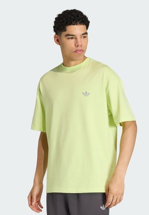Junger Mann, der ein hellgrünes Adidas T-Shirt und dunkle Shorts trägt und zur Seite gegen einen einfarbigen hellen Hintergrund schaut.
