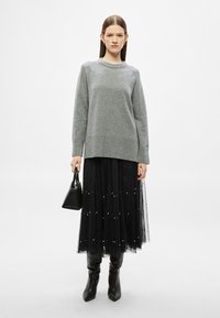 Γκρι πλεκτό πουλόβερ με στρογγυλή λαιμόκοψη, συνδυασμένο με μαύρη φούστα από τούλι διακοσμημένη με λευκές πέρλες. Το μοντέλο κρατά μια μικρή μαύρη τσάντα χειρός.