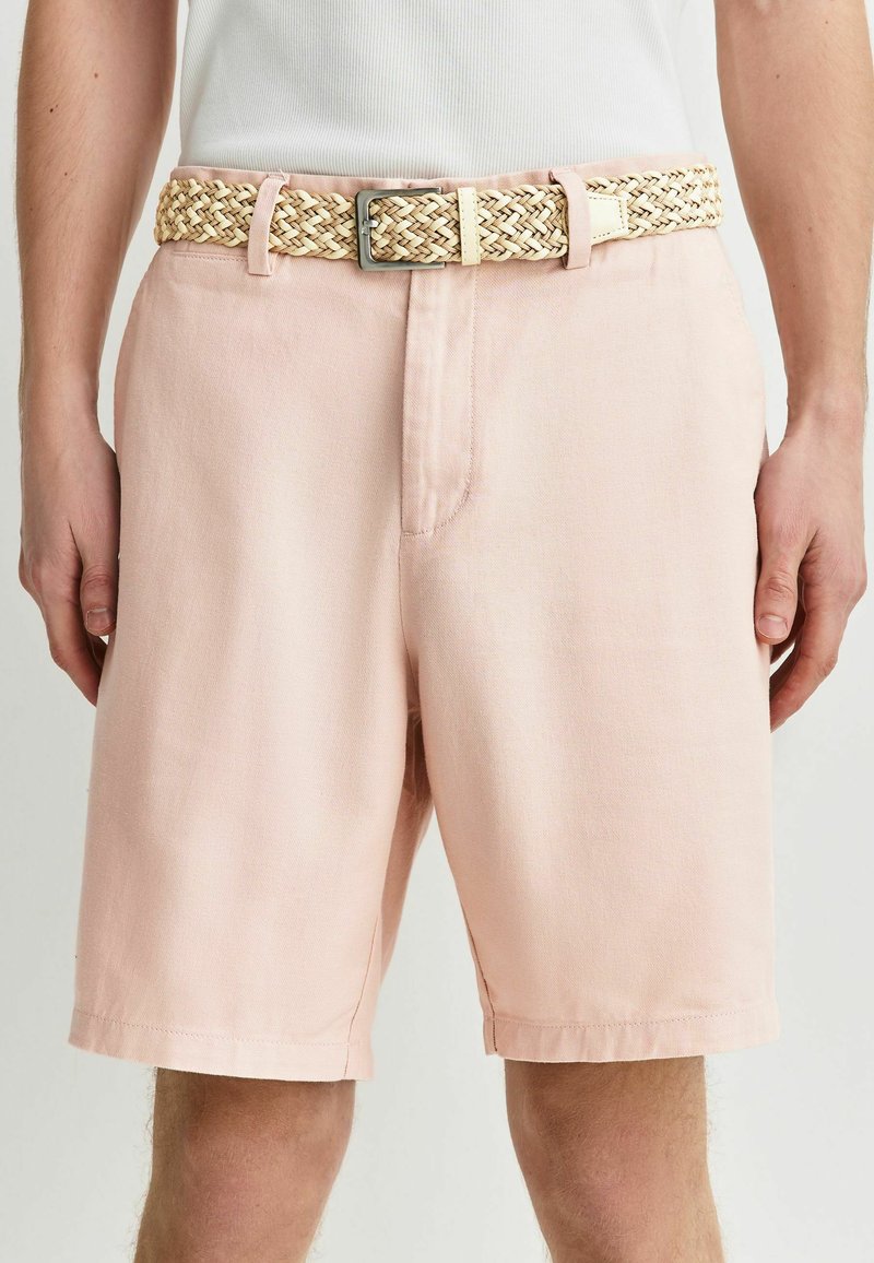 Shorts rose clair longueur genou avec une ceinture beige tissée et une boucle argentée, portés avec un top blanc sans manches.